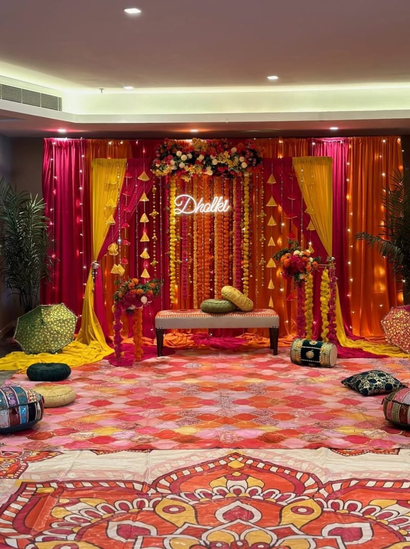 Dholki stage with pink and orange drapes, marigold strings and neon sign – Events In Sialkot